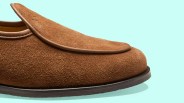 Brown suede loafer with leather trim and black sole on a light blue background.