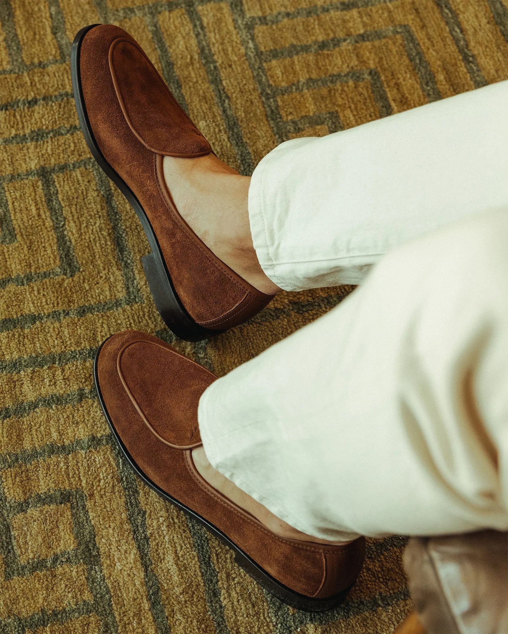Pair of brown suede loafers worn with off-white pants on a patterned carpet.