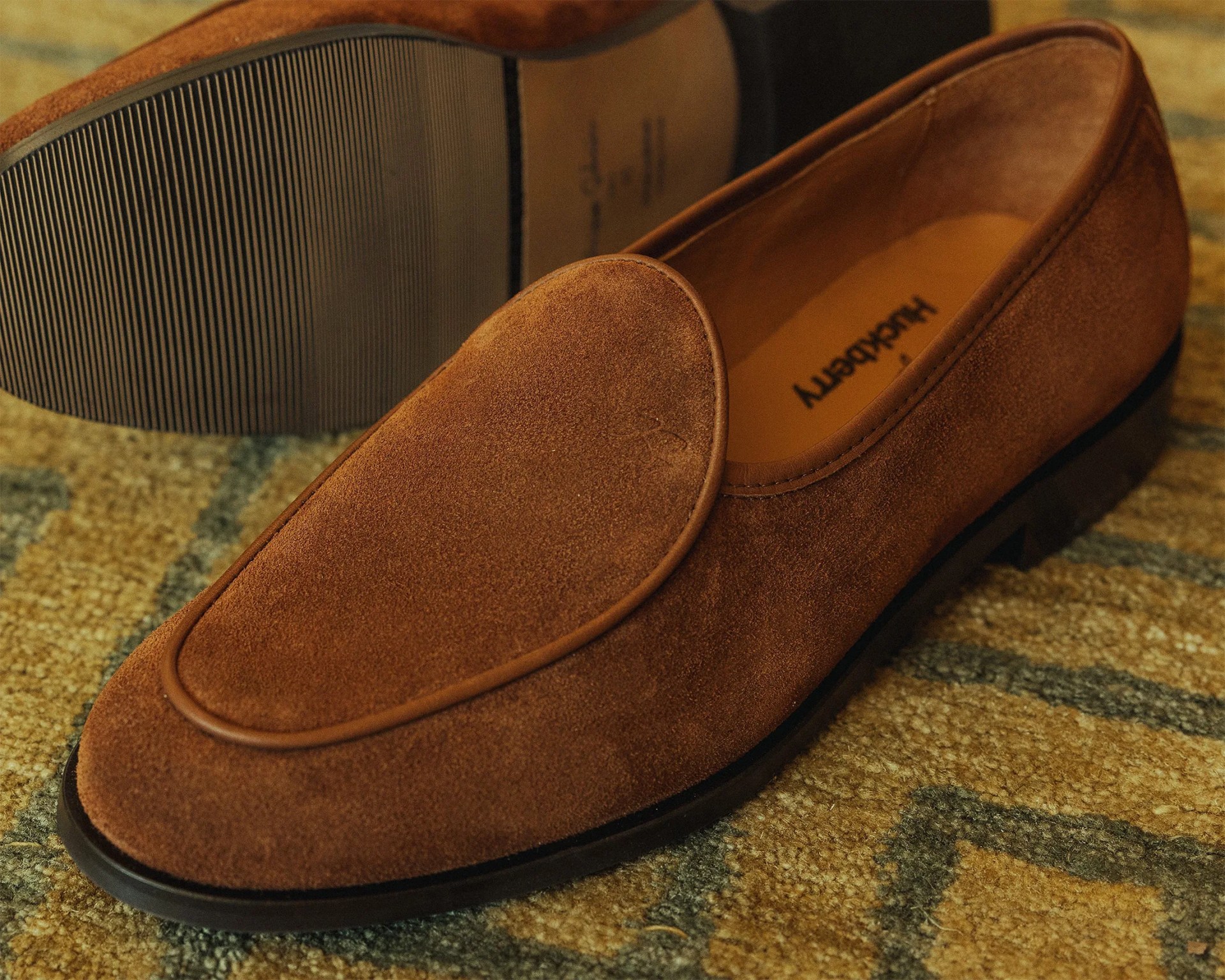 Pair of brown suede loafers with black soles on a patterned carpet.