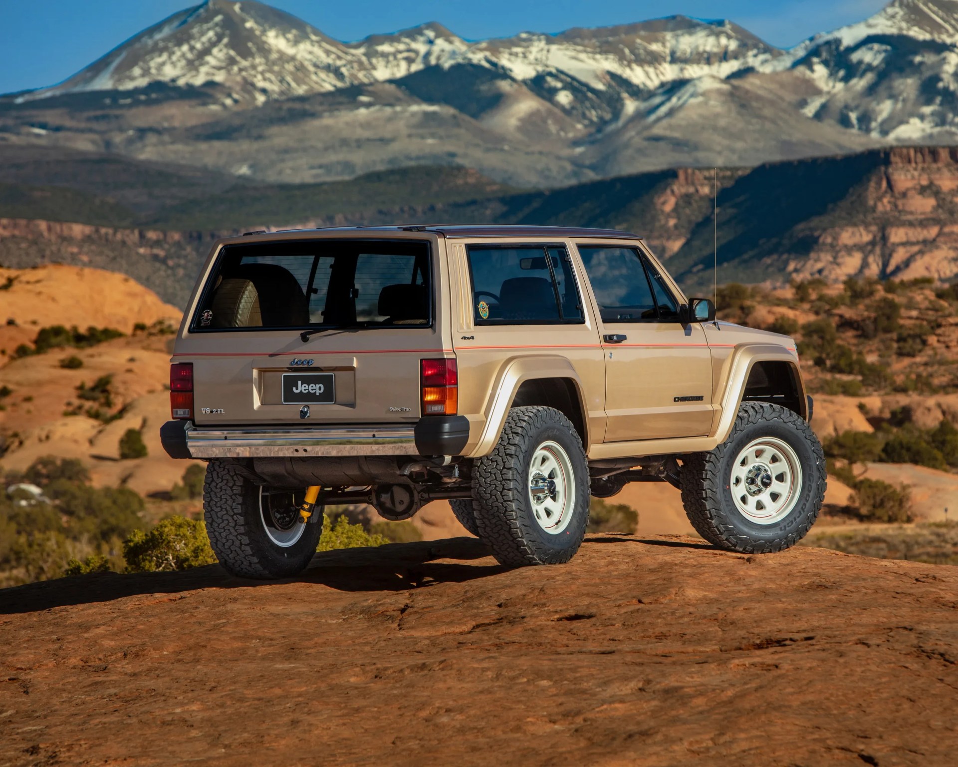 Beige Jeep Cherokee with off-road tires parked on rocky terrain with snow-capped mountains in the background.