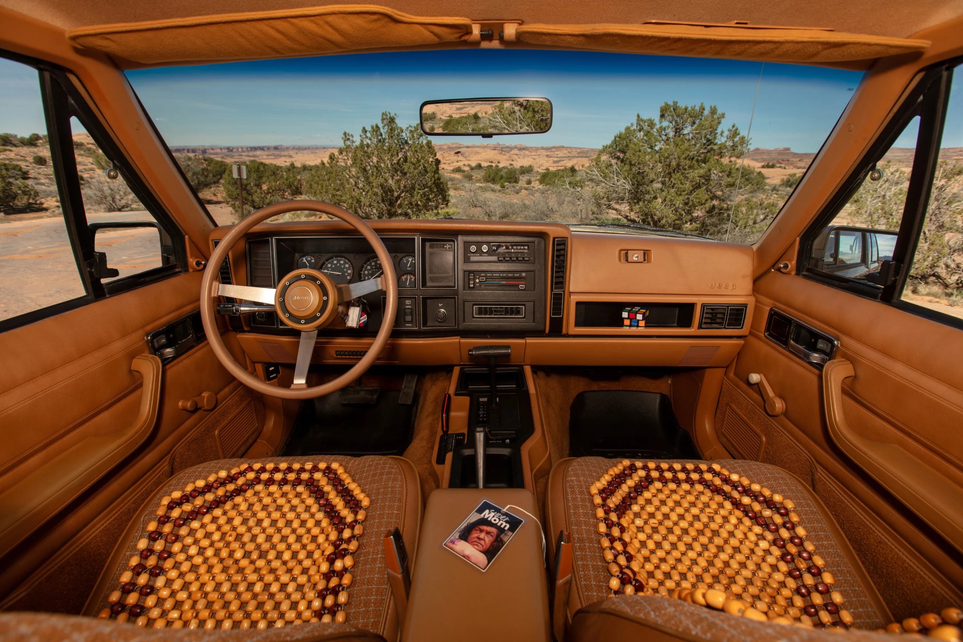 Interior of a vintage tan Jeep with wooden bead seat covers, a Rubik's cube on the dashboard, and a "Wild Mom" air freshener on the center console.