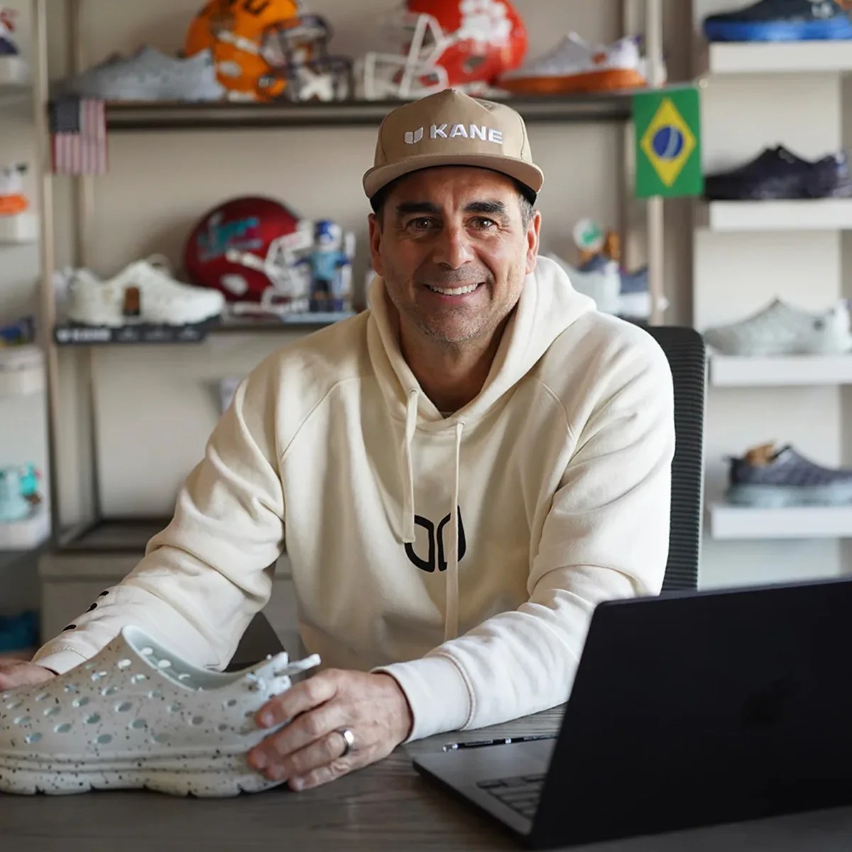 Man in beige hoodie and tan cap holding a white speckled sneaker at a desk with a laptop.