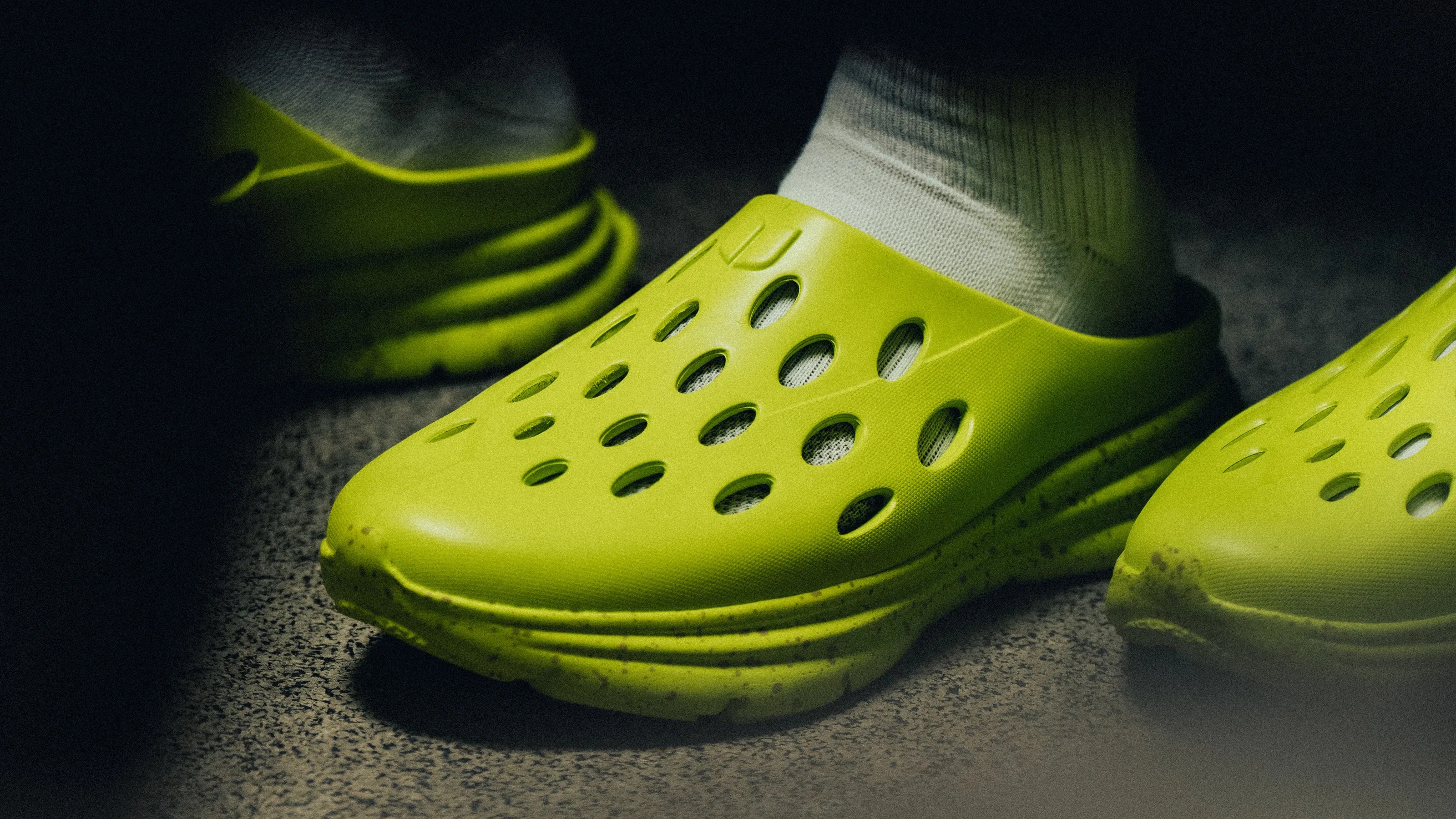 Bright yellow slip-on shoes with oval ventilation holes worn with white socks on a textured floor.