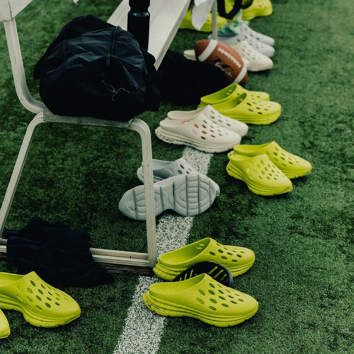 Pairs of yellow and white slip-on clogs arranged on artificial turf near a white bench with a black bag and a football.