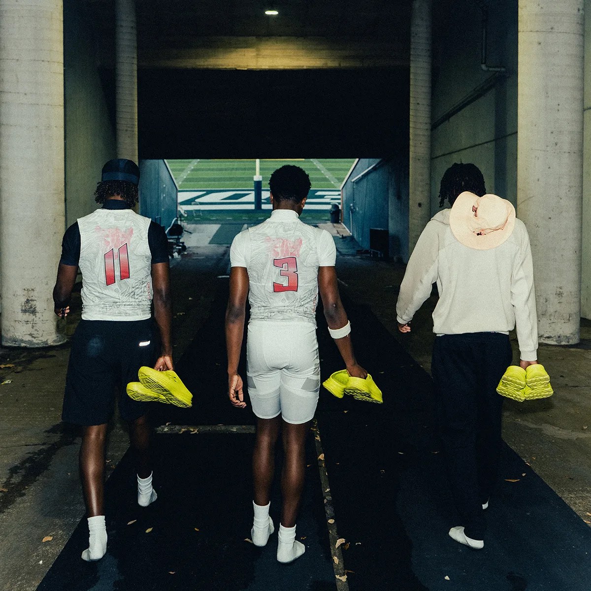 Three athletes walking away in a tunnel holding bright yellow cleats, two wearing white sports uniforms with numbers 11 and 3.