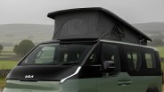 Green camper van with black pop-up roof tent and black front hood parked in a grassy field.