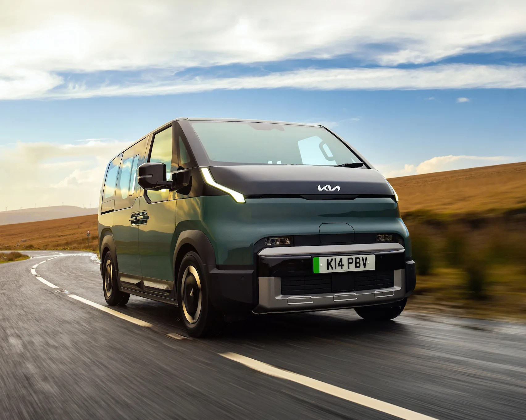 Green and black Kia electric van driving on a wet road with a cloudy sky and brown hills in the background.