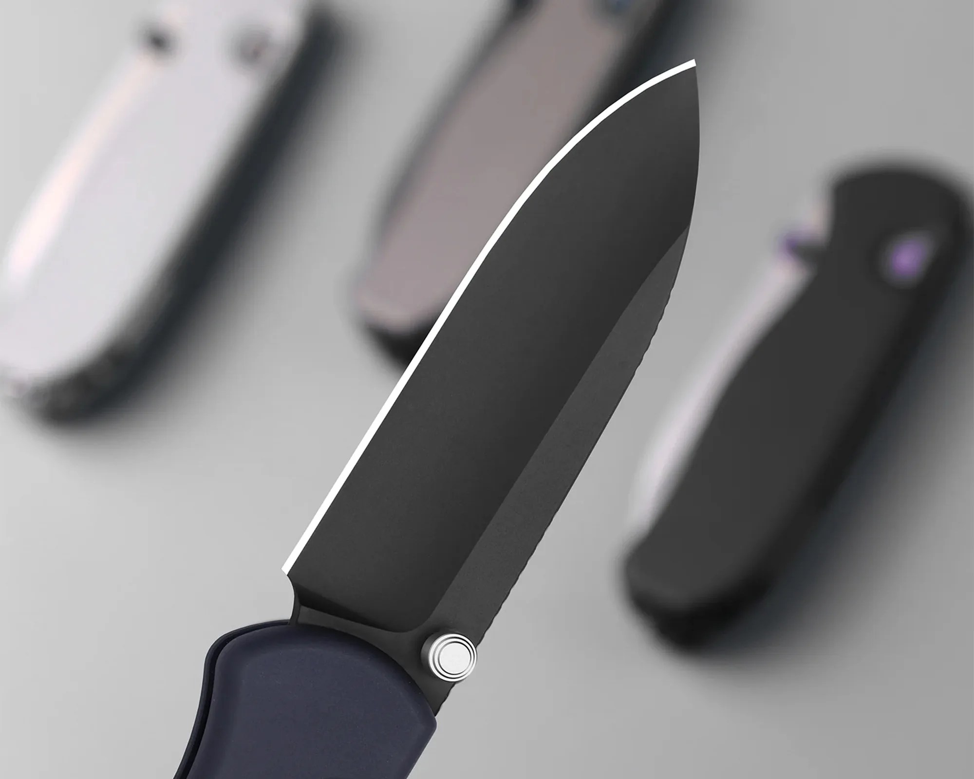 Close-up of a black folding knife blade with a dark blue handle and three blurred knives in the background.