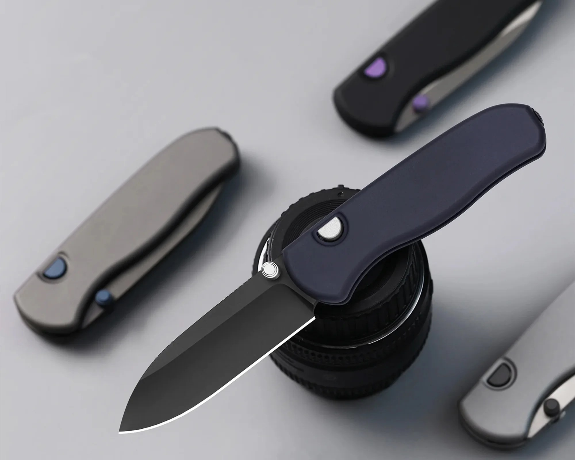 Black folding knife with a dark blue handle resting on a camera lens, surrounded by three closed knives with gray and black handles.