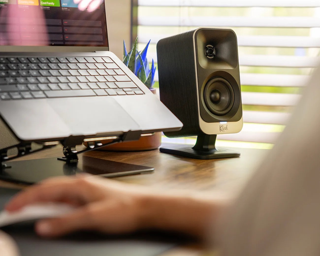 Black Klipsch speaker with silver front panel on a wooden desk next to a laptop and a small potted plant.