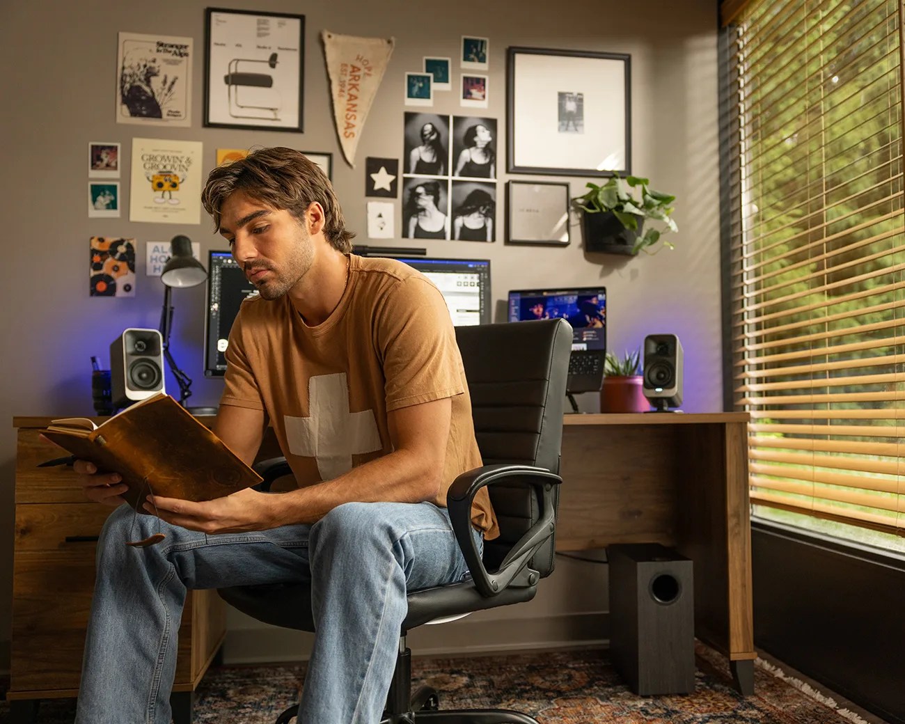 Person in a brown t-shirt with a white cross reading a book while sitting on a black office chair in a home office.