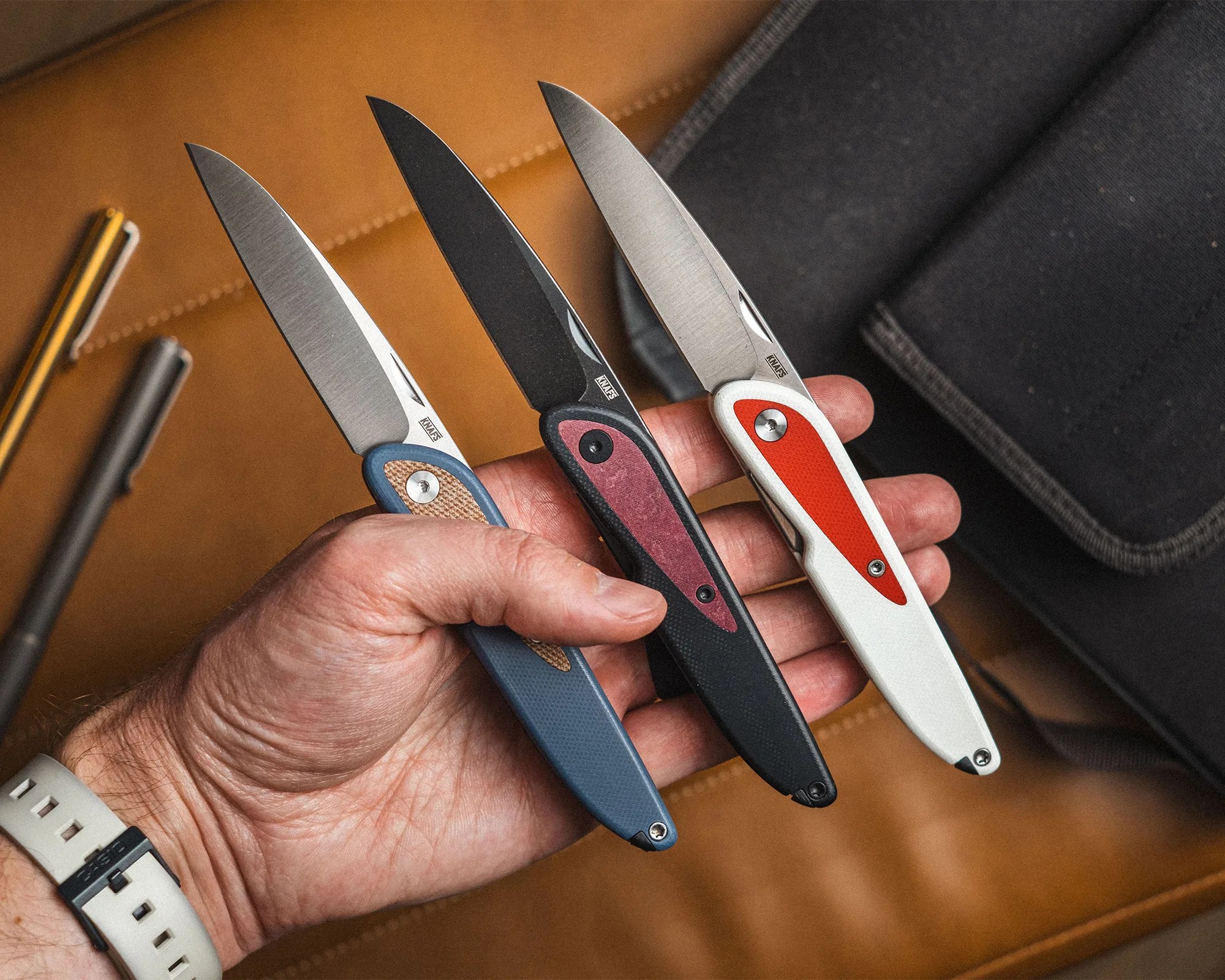 Three folding knives with stainless steel blades and colored handles (blue with tan, black with red, white with orange) held in a hand over a brown leather surface.