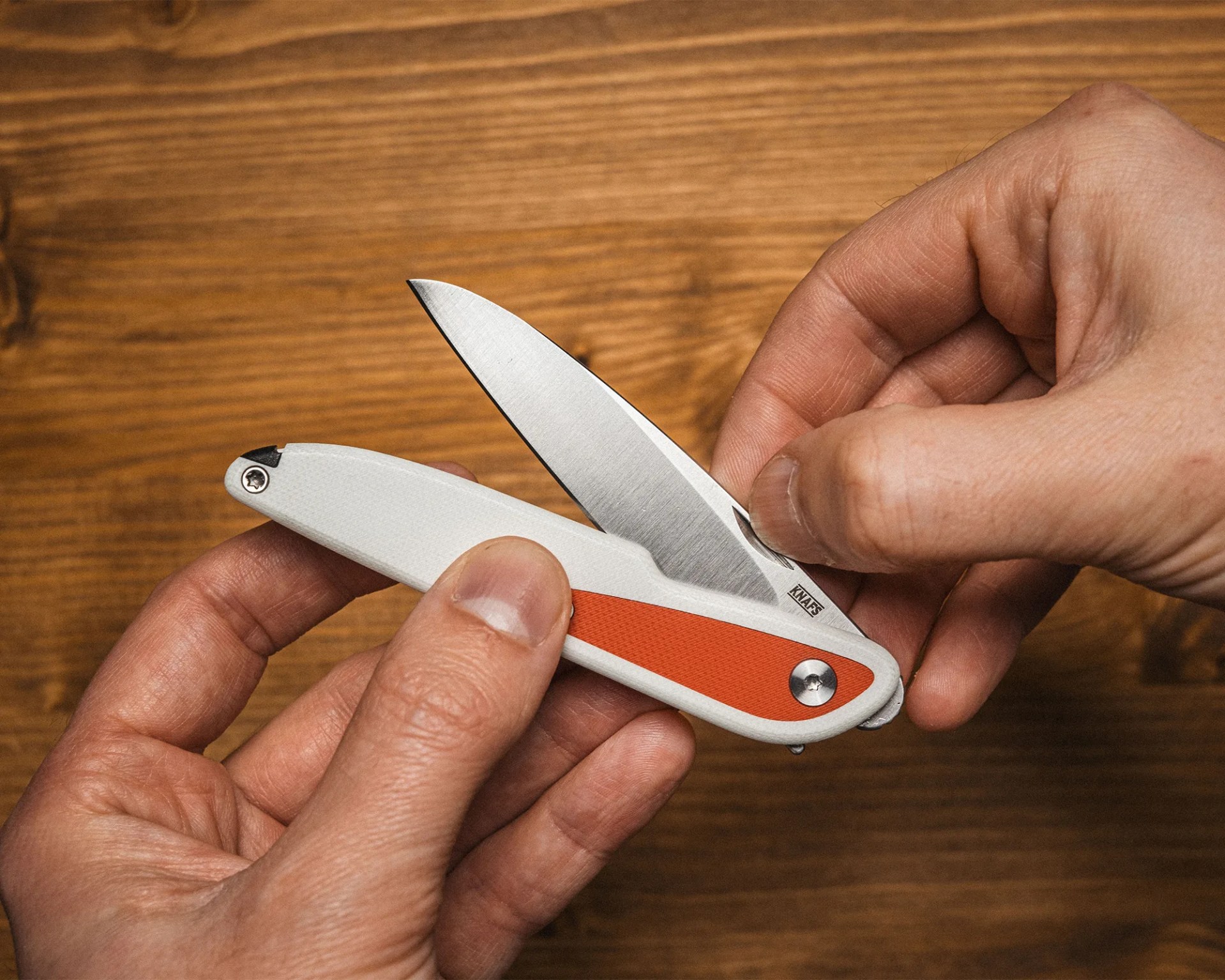 Hands holding a folding pocket knife with a white and orange handle and a partially opened stainless steel blade.