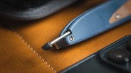 Blue folding knife with a metal clip and a wooden inlay handle resting on brown leather next to a smartphone camera.