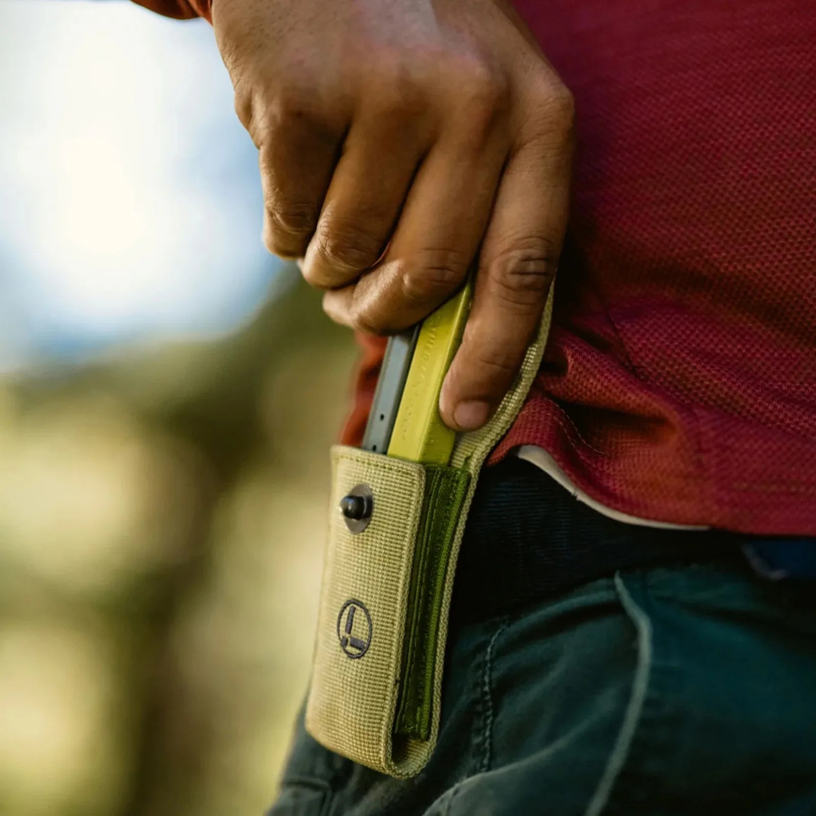 Hand placing a yellow and gray magazine into a beige fabric pouch attached to a belt on green pants.