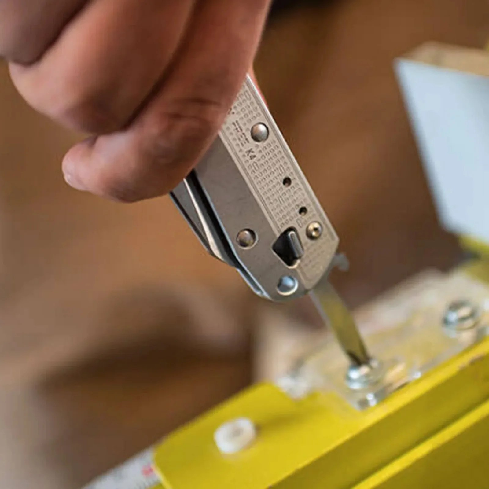 Hand using a silver multi-tool with a flathead screwdriver to tighten a screw on a yellow object.