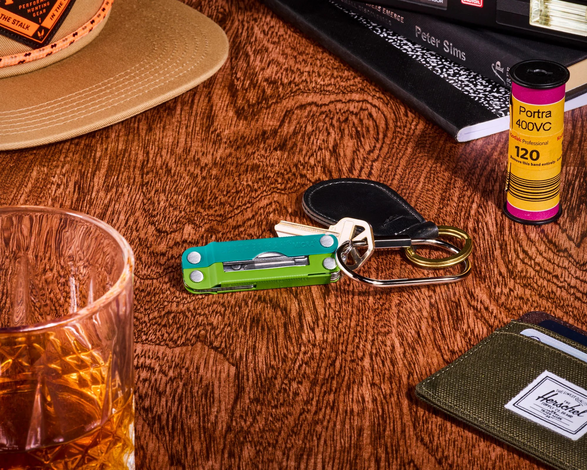 Set of keys with a green and blue multi-tool on a wooden surface next to a glass of amber liquid, a tan cap, a film canister, and a green wallet.