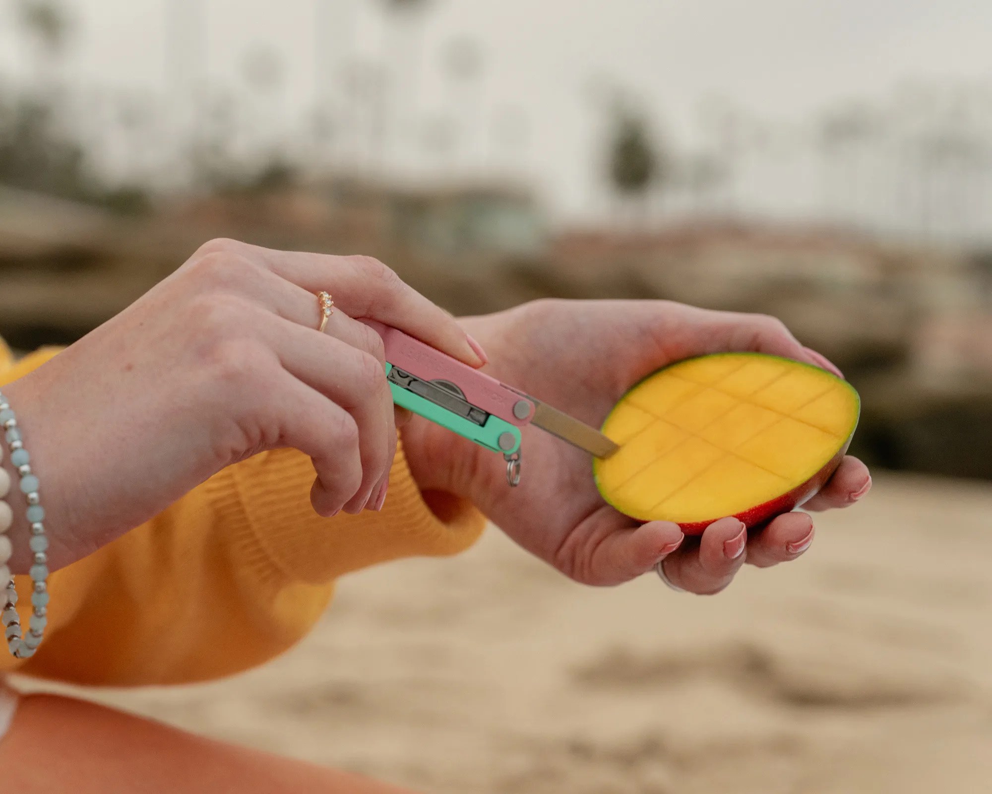 Hands holding a halved mango with a pink and green multi-tool knife cutting grid lines into the fruit.