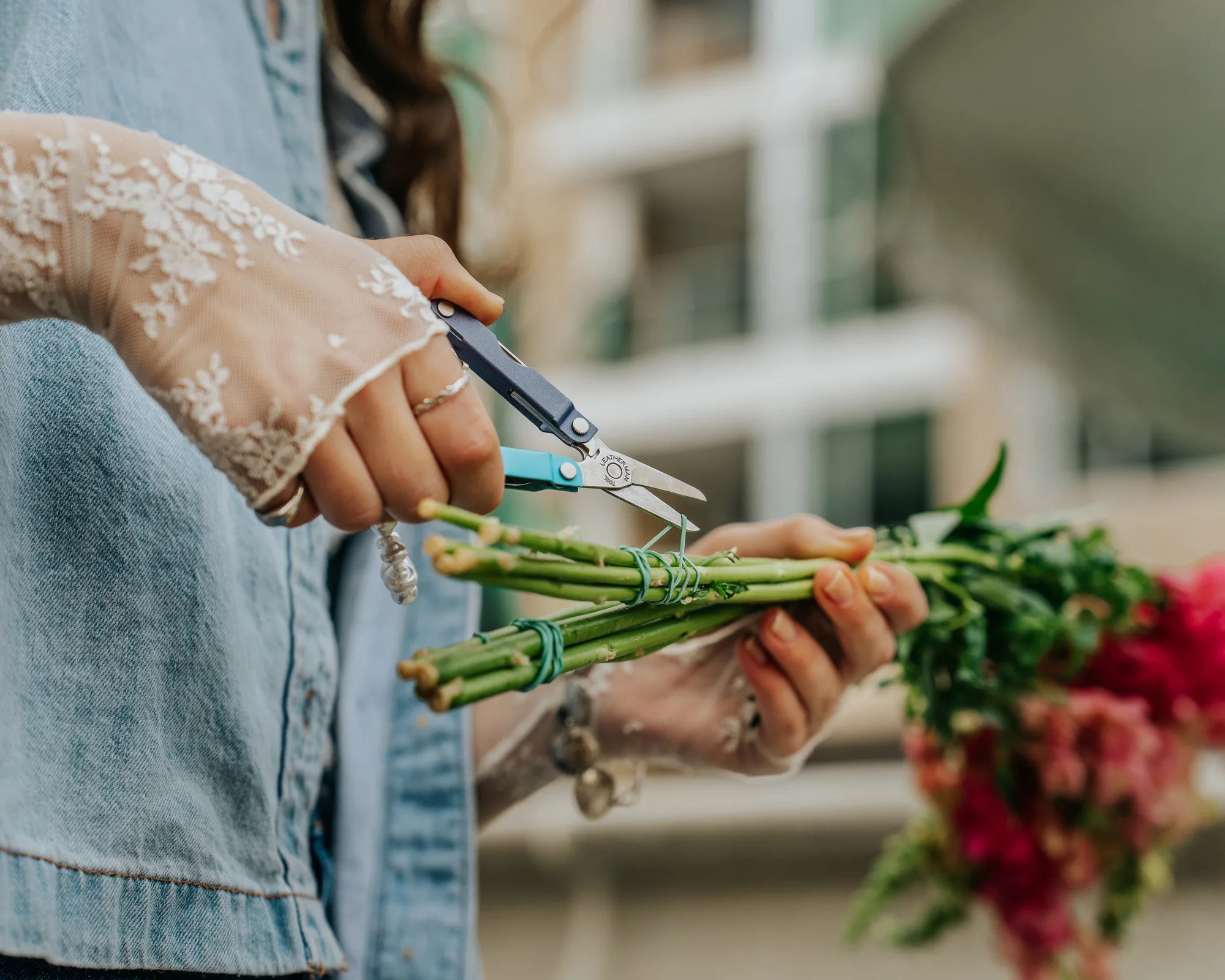 Hands wearing lace gloves and denim jacket using small blue scissors to trim green flower stems.