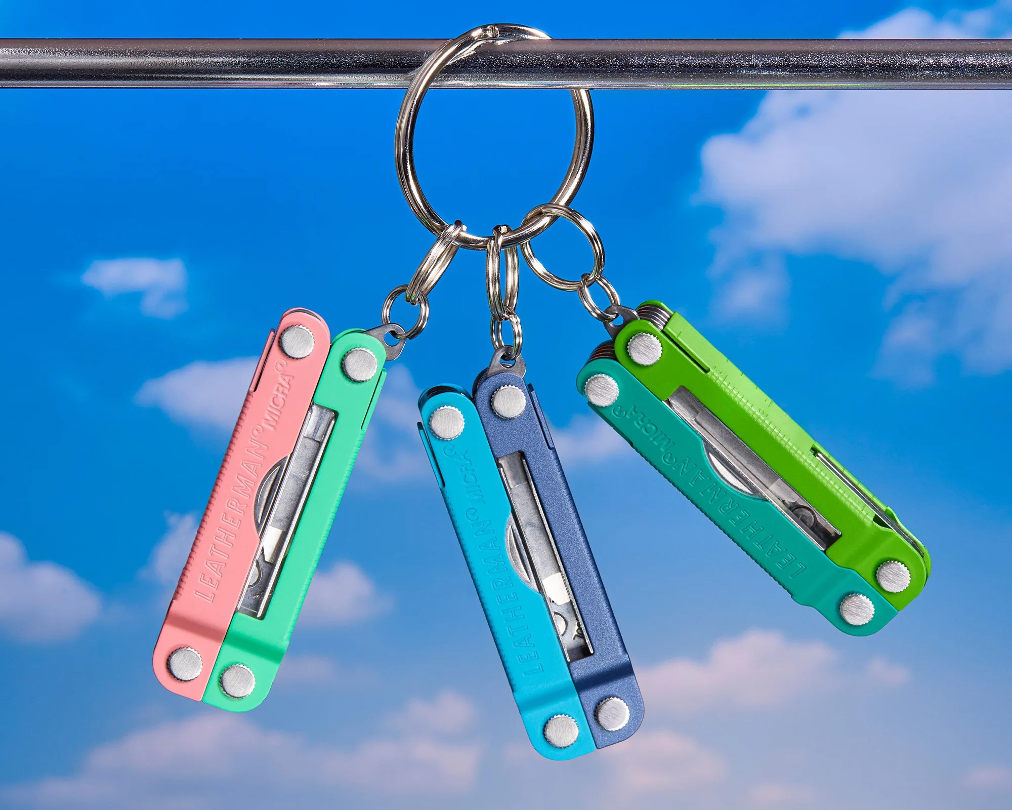 Three colorful Leatherman Micra multi-tools in pink/green, blue/navy, and green/teal hanging on a metal ring against a blue sky with clouds.