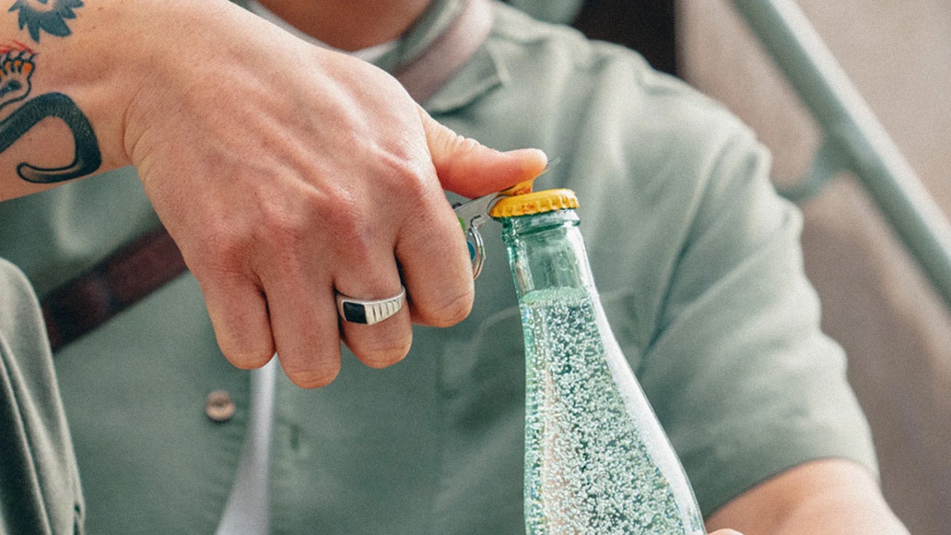 Hand with a silver ring and tattoo using a bottle opener to open a yellow-capped sparkling water bottle.