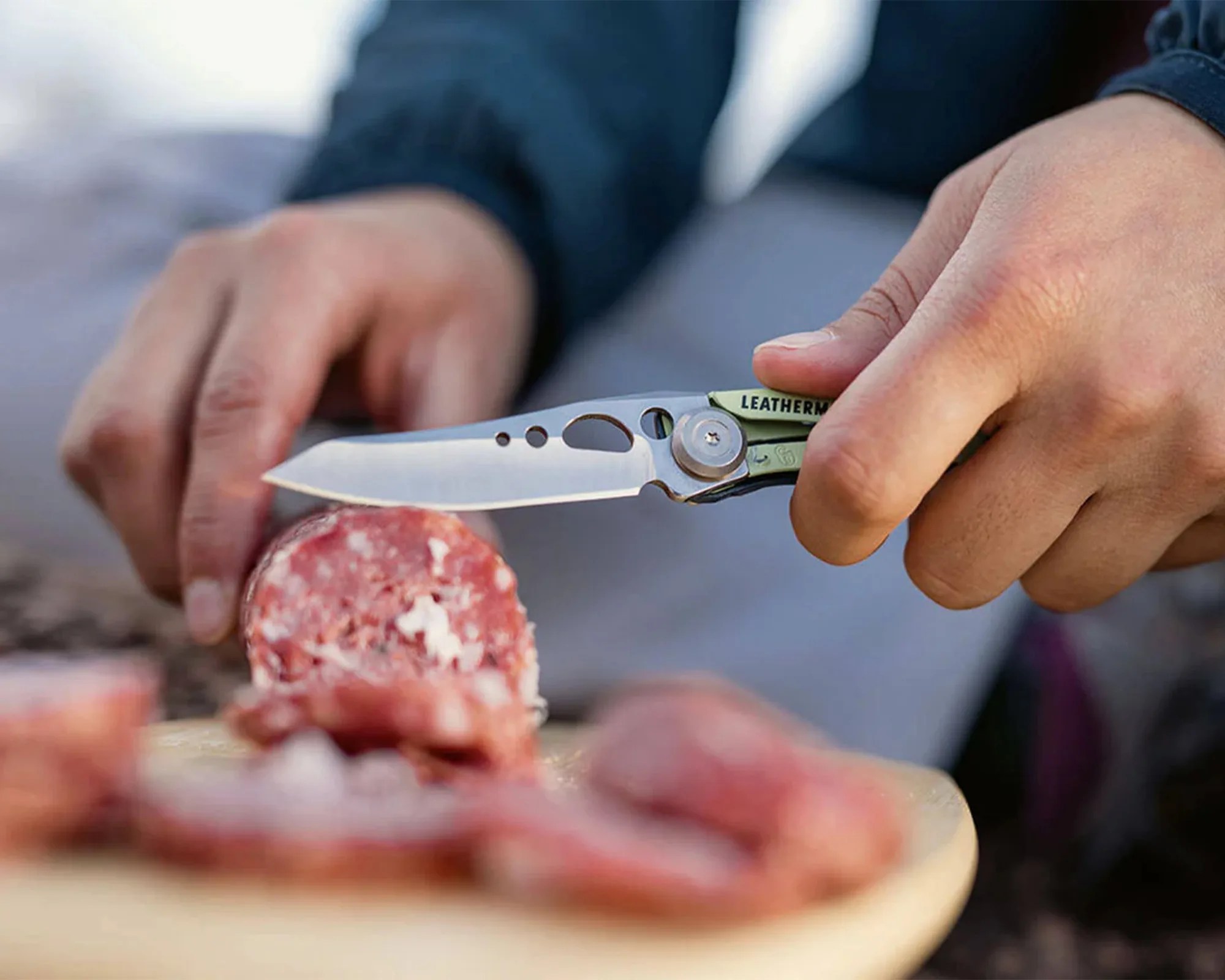 Hand holding a Leatherman folding knife slicing a piece of salami on a wooden board.