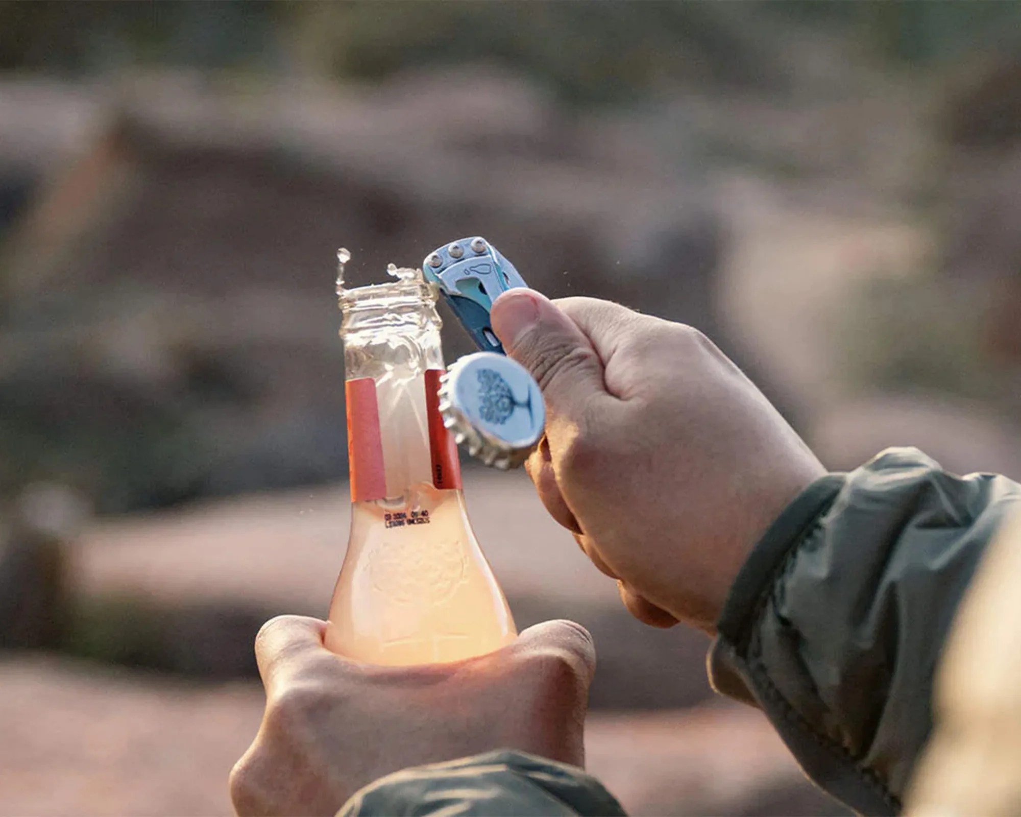 Hands opening a glass bottle with a blue multi-tool bottle opener outdoors.