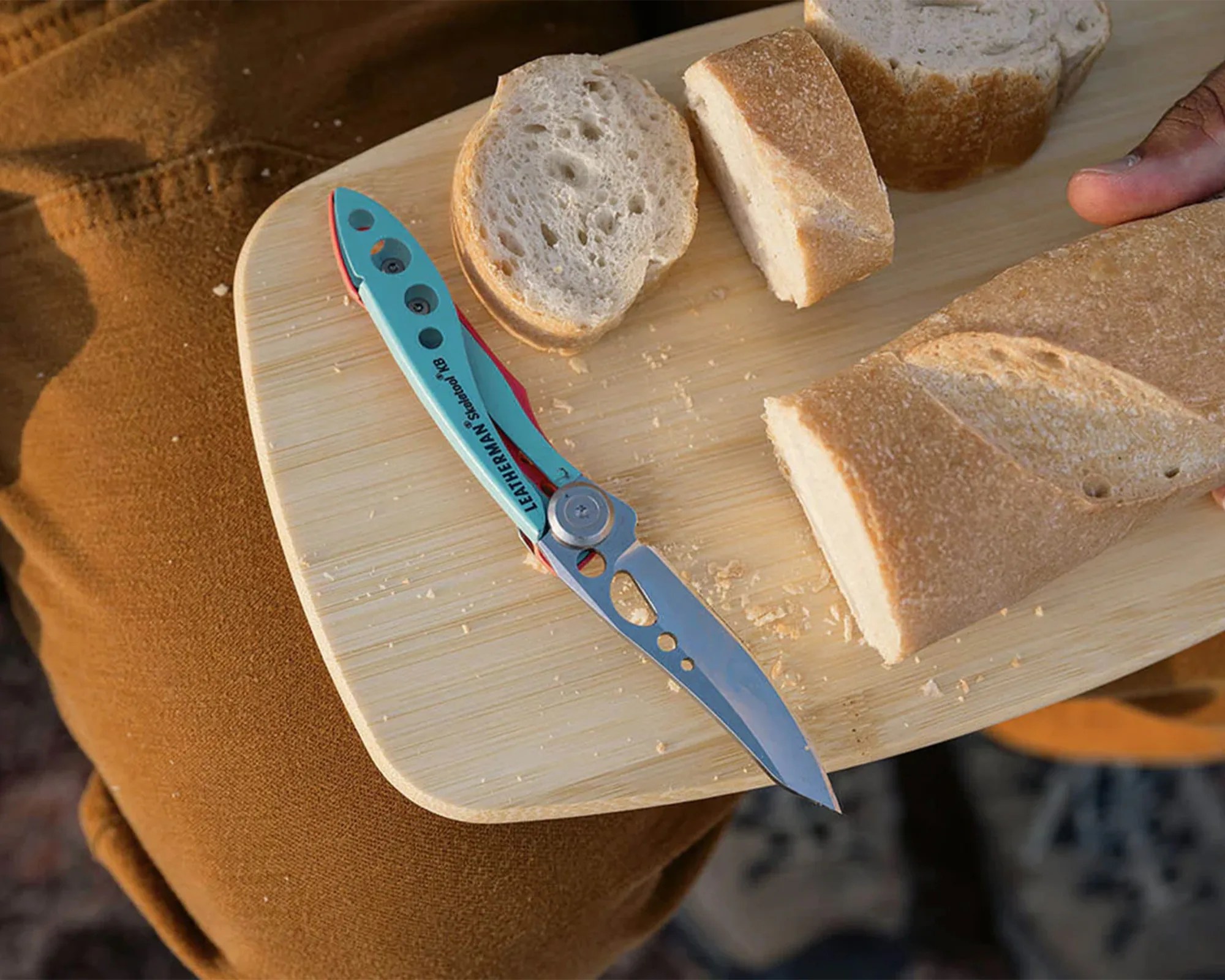 Light blue Leatherman Skeletool BK folding knife with a partially serrated blade on a wooden cutting board with sliced bread.