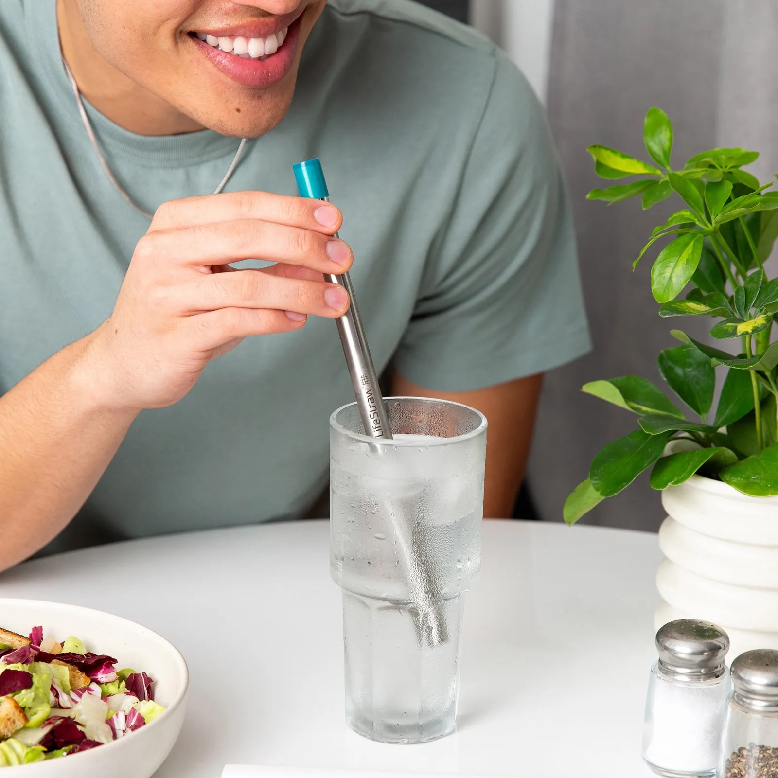 Person holding a stainless steel LifeStraw in a tall glass of ice water on a white table with a salad bowl and potted plant nearby.
