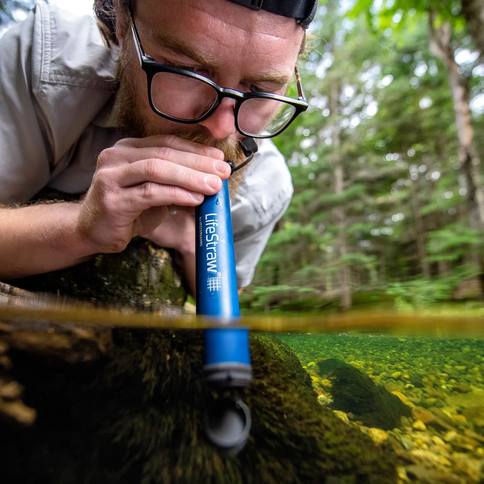 Person using a blue LifeStraw water filter to drink from a clear forest stream.