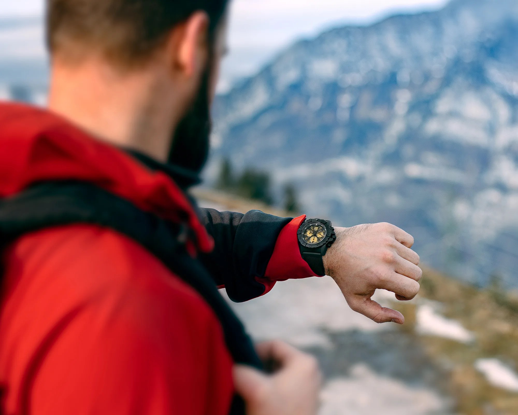Black and gold wristwatch worn by a person in a red jacket with a mountainous background.