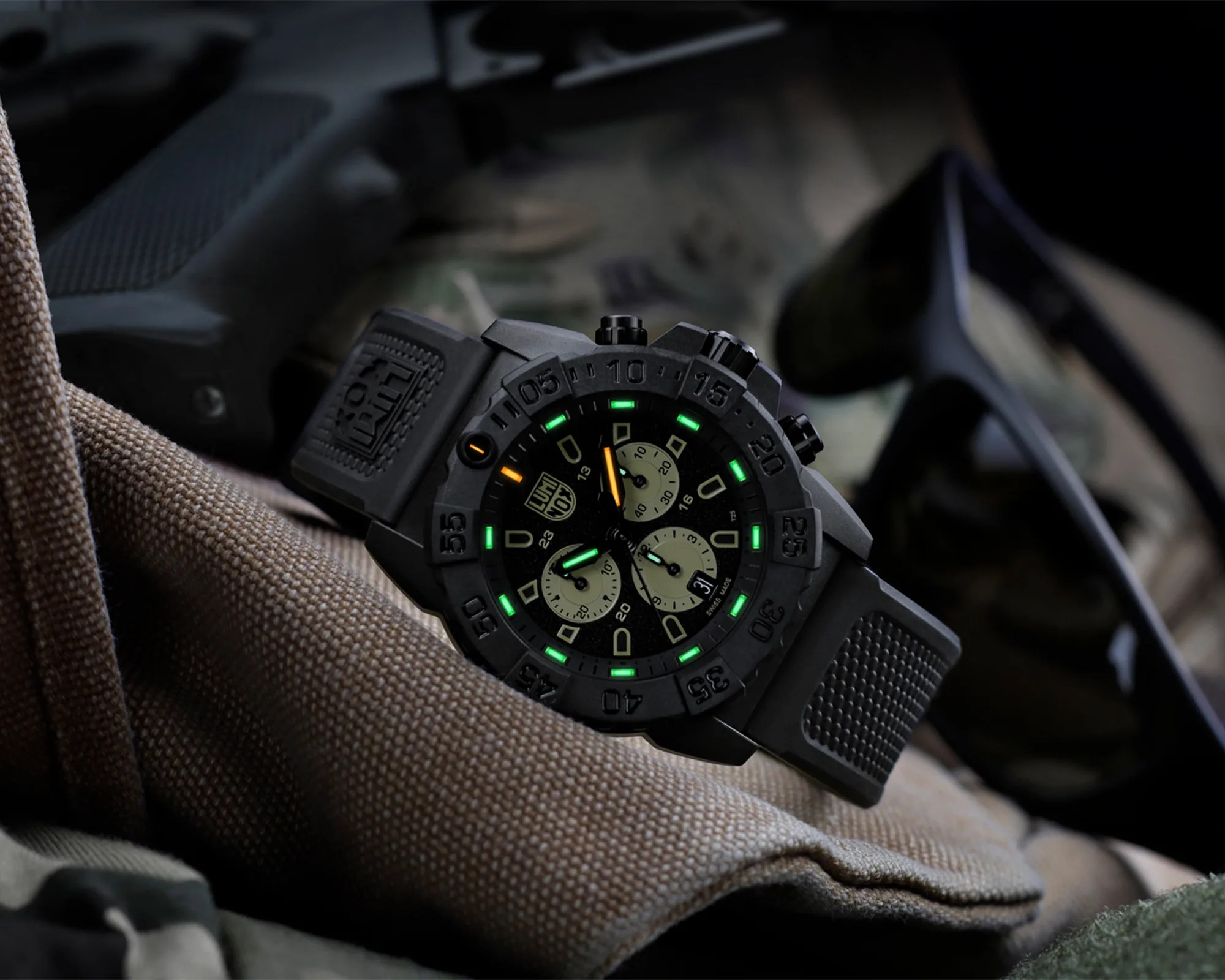 Black tactical wristwatch with green and orange luminescent markers on a textured fabric surface.