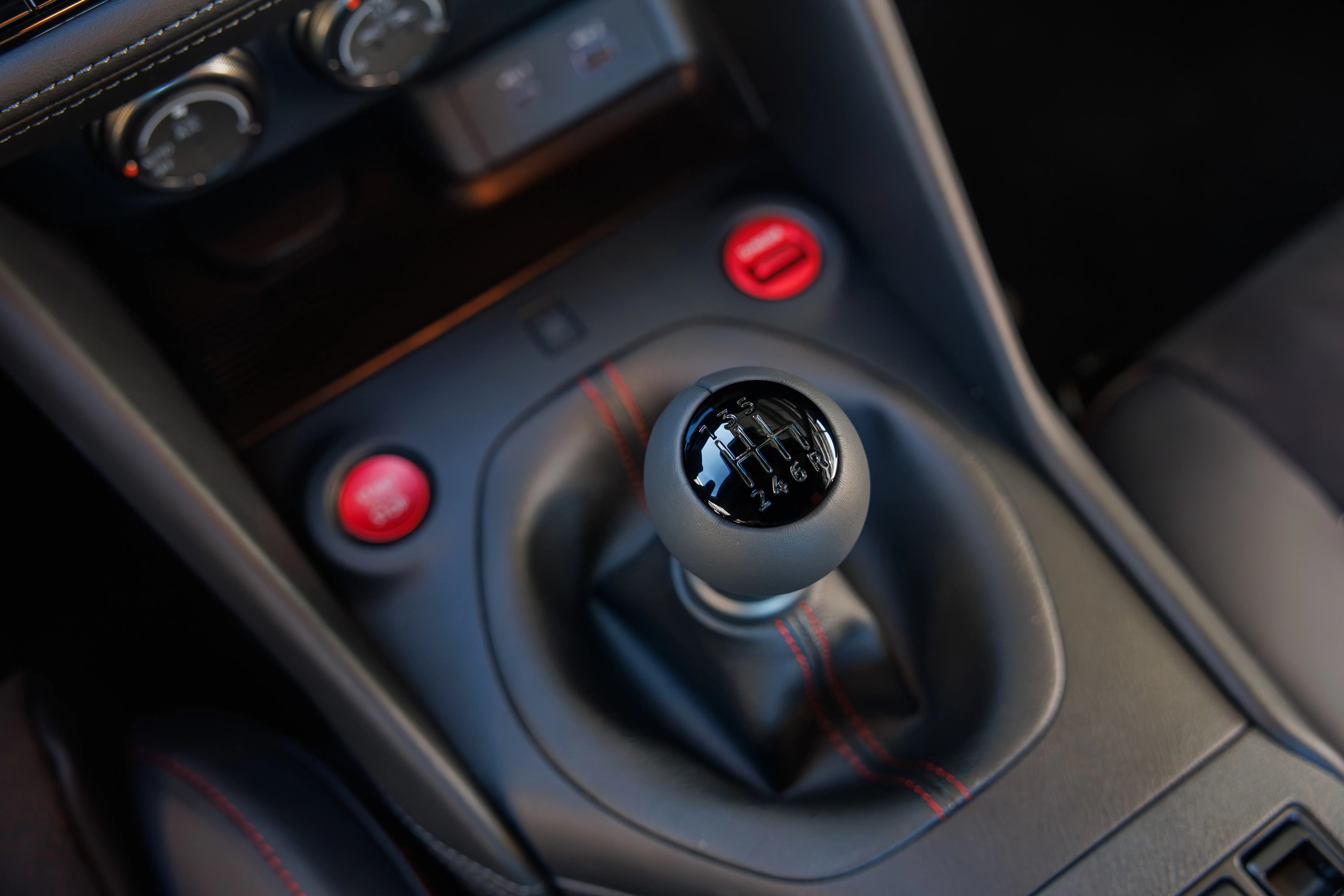 Black manual gear shift knob with six-speed pattern and reverse, surrounded by black leather with red stitching in a car interior.