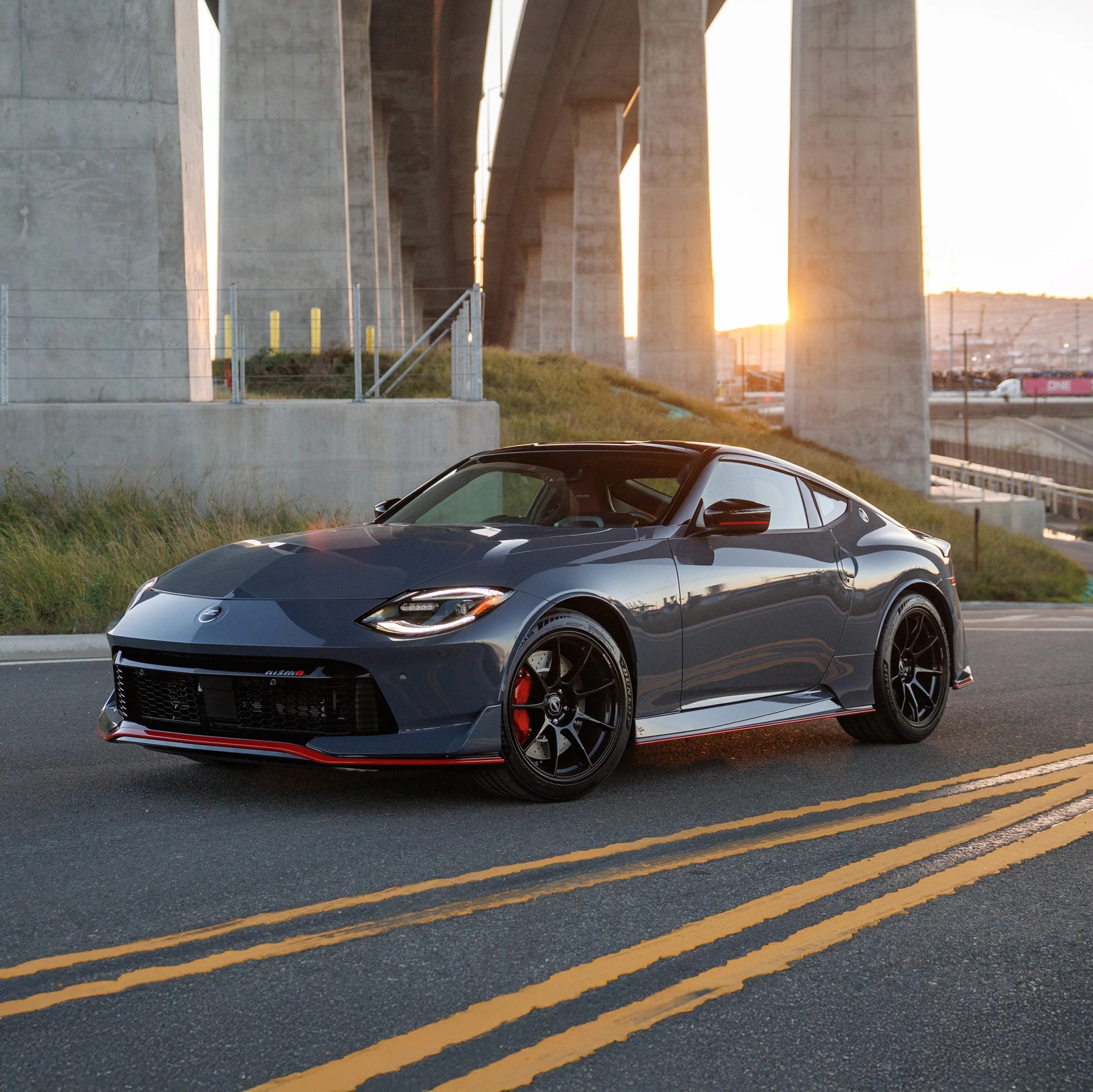 Dark gray Nissan sports car with black wheels and red brake calipers parked on a road at sunset under a concrete bridge.