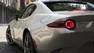 Rear side view of a silver Mazda MX-5 with illuminated taillight parked on a city street.