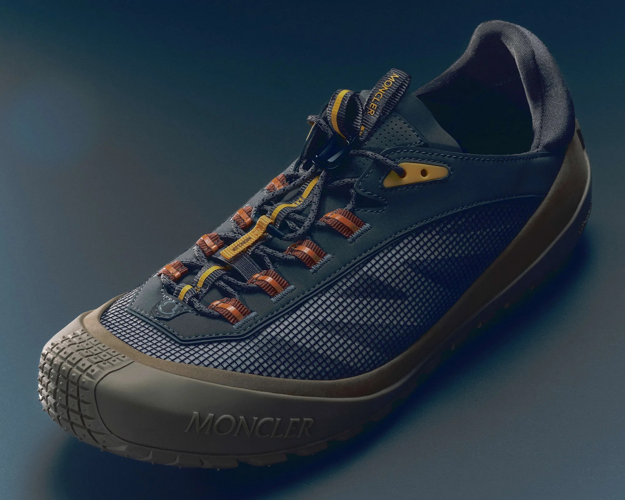 Dark gray Moncler sneaker with mesh sides, brown and yellow lace loops, and textured rubber sole.