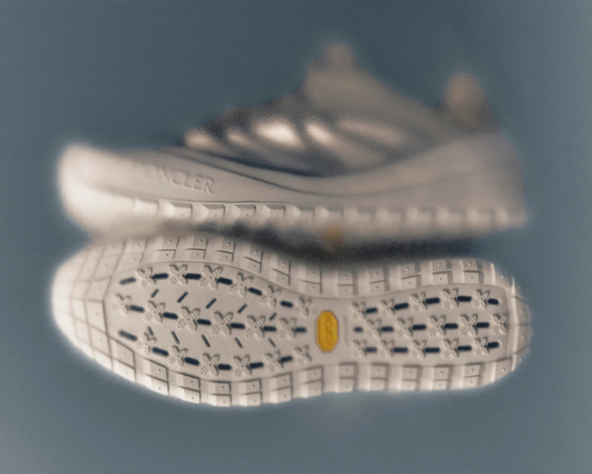 Pair of white sneakers with textured soles, one showing the side with "MONCLER" branding and the other showing the sole with a yellow Vibram logo.