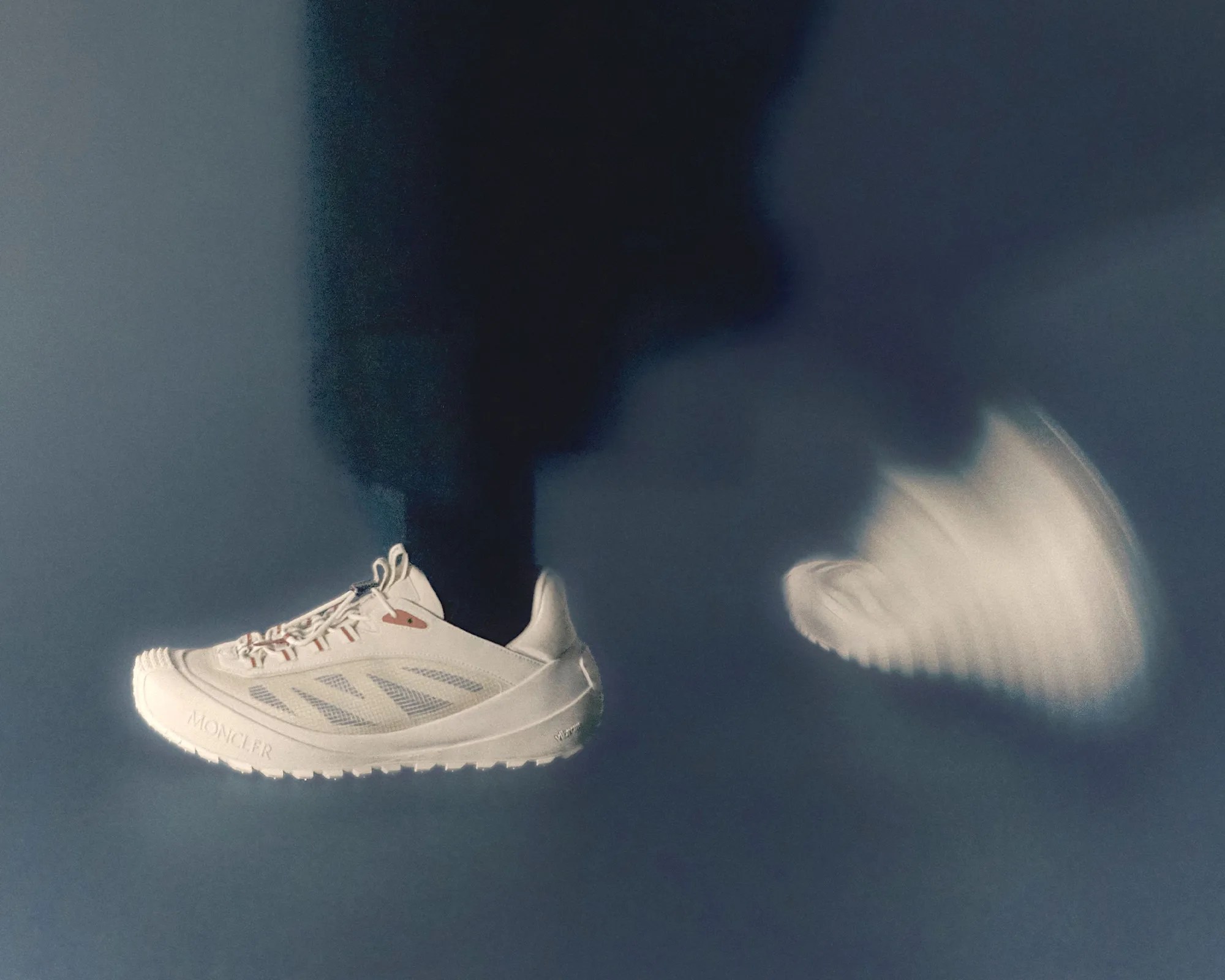 White Moncler sneaker with mesh side panels and textured sole worn by a person in motion against a dark background.