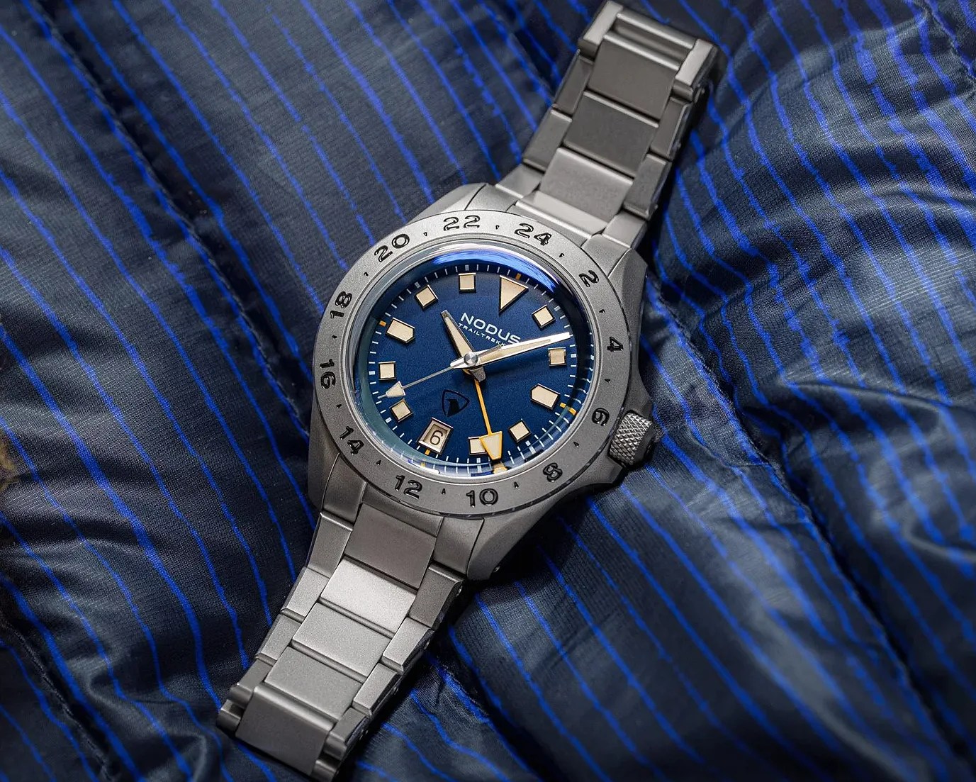 Matte silver metal wristwatch with blue dial, beige hour markers, date window, and 24-hour bezel on blue striped fabric.