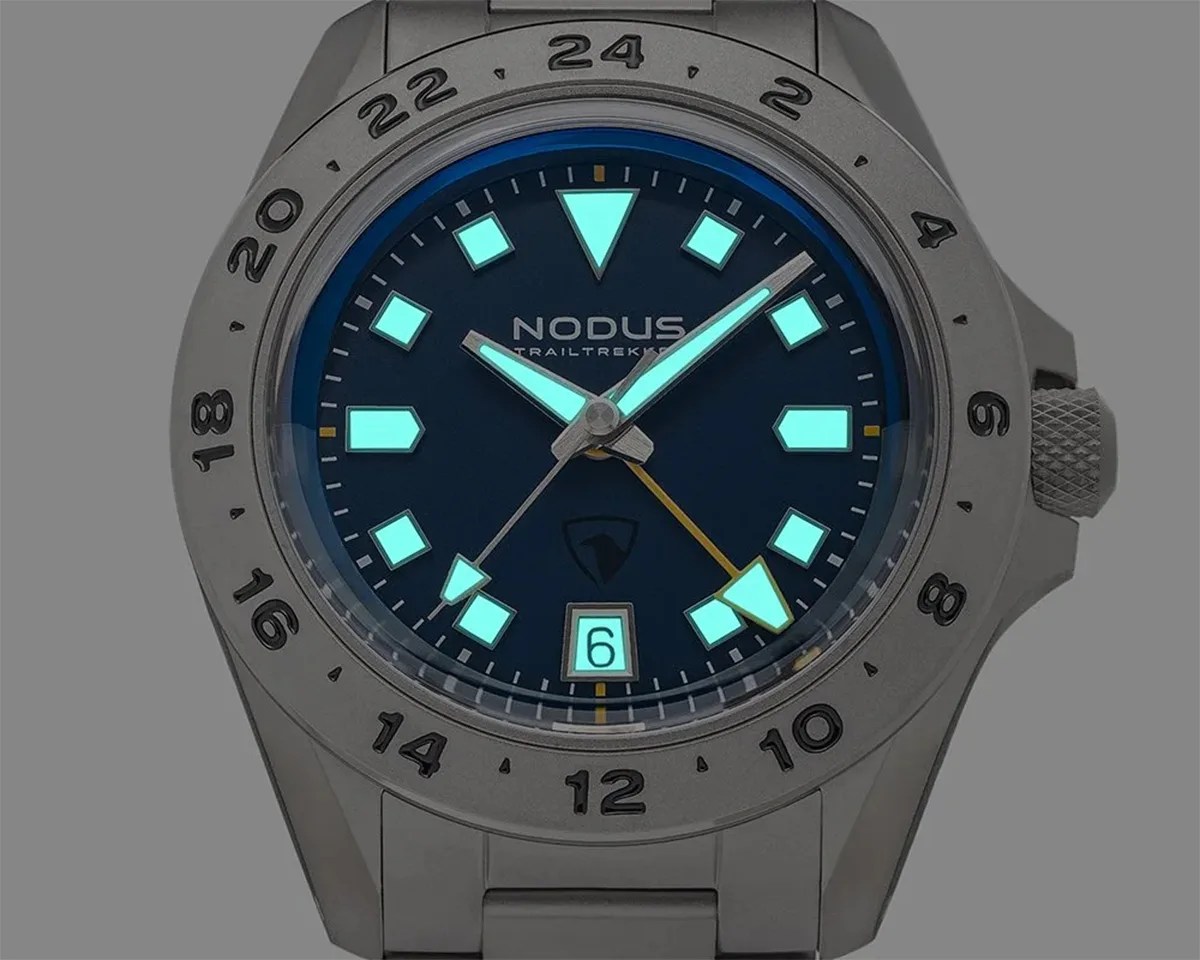 Close-up of a Nodus Trailkeeper watch with luminescent hour markers and hands on a dark dial, stainless steel case and bracelet.