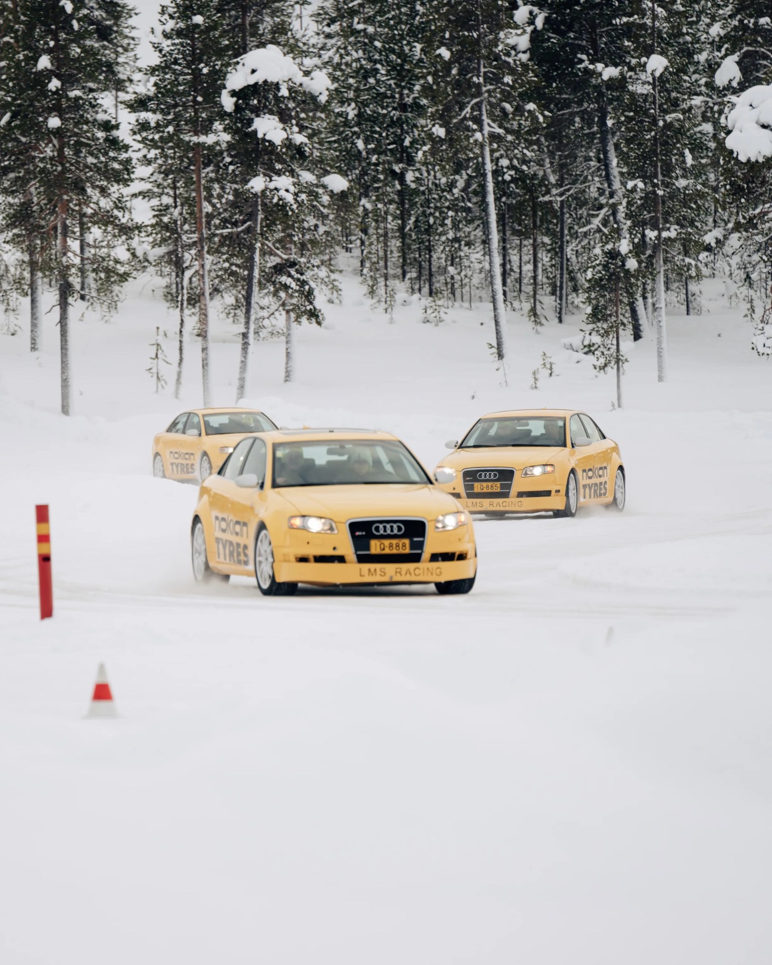 Three yellow Audi LMS Racing cars with Nokian Tyres branding driving on a snowy track surrounded by snow-covered trees.