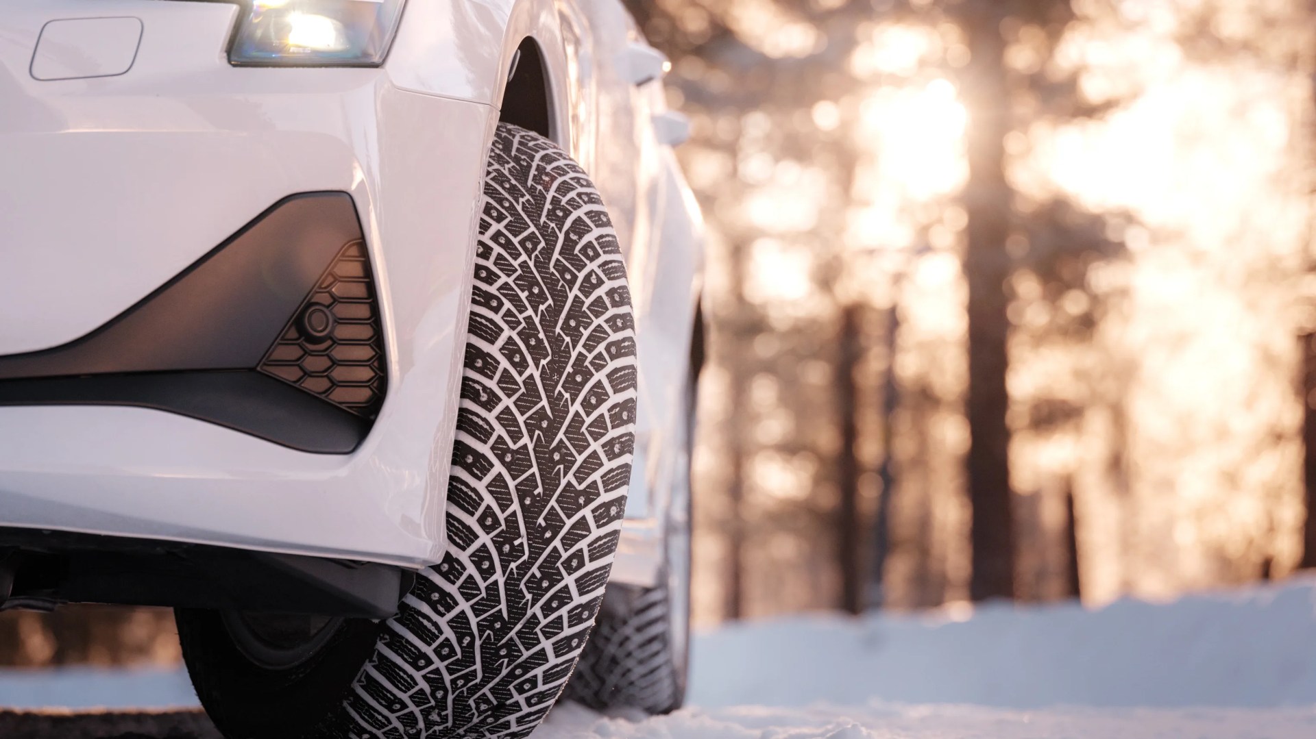 Close-up of a white car's front tire with winter tread pattern on snow-covered ground at sunset.