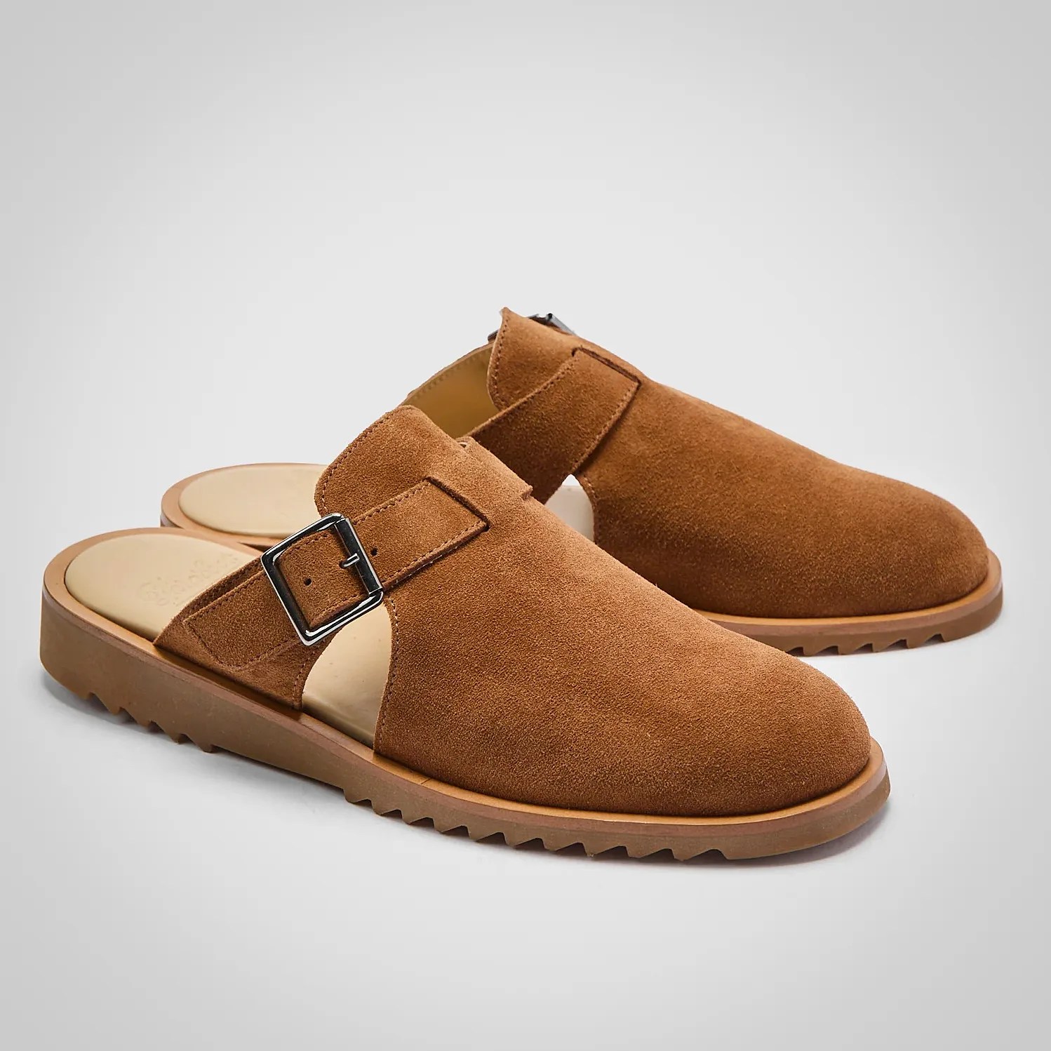 Pair of brown suede slip-on shoes with a silver buckle and ridged rubber soles on a gray background.