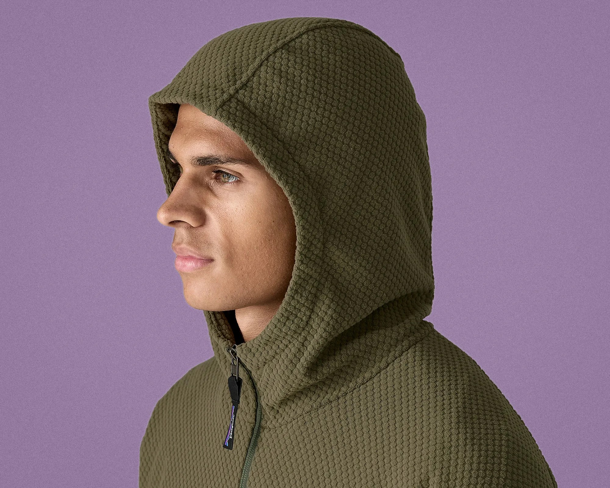 Man wearing an olive green textured hooded jacket with a zipper against a purple background.