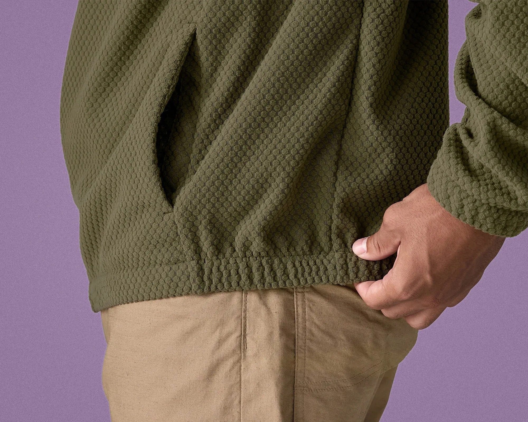 Close-up of a hand adjusting the hem of an olive green textured jacket worn over beige pants.
