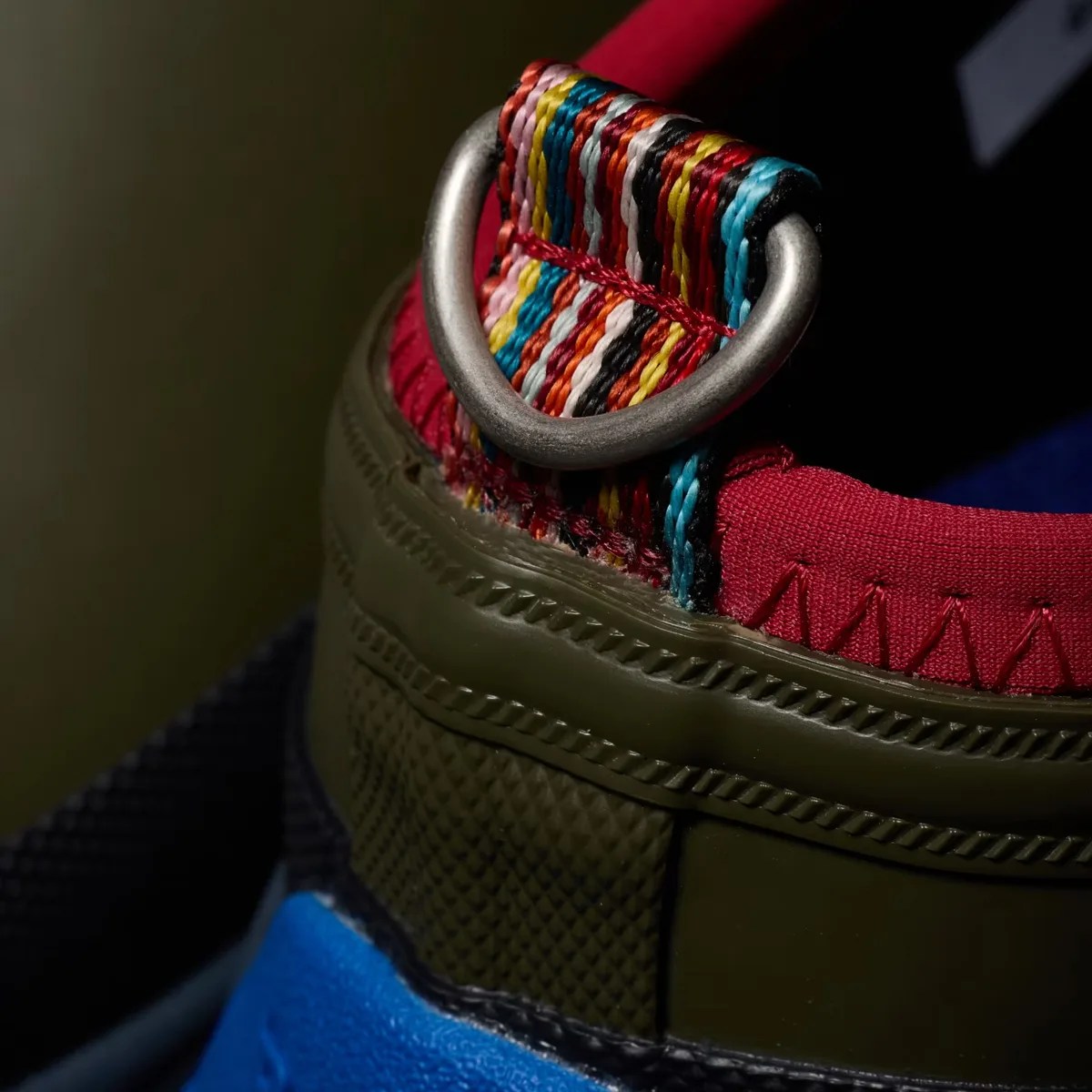 Close-up of a sneaker heel with multicolored woven pull tab and metal D-ring, featuring red fabric and olive green and blue sole.
