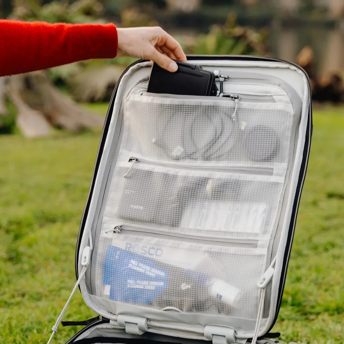 Hand placing a black electronic device into a white mesh zippered compartment inside an open suitcase.