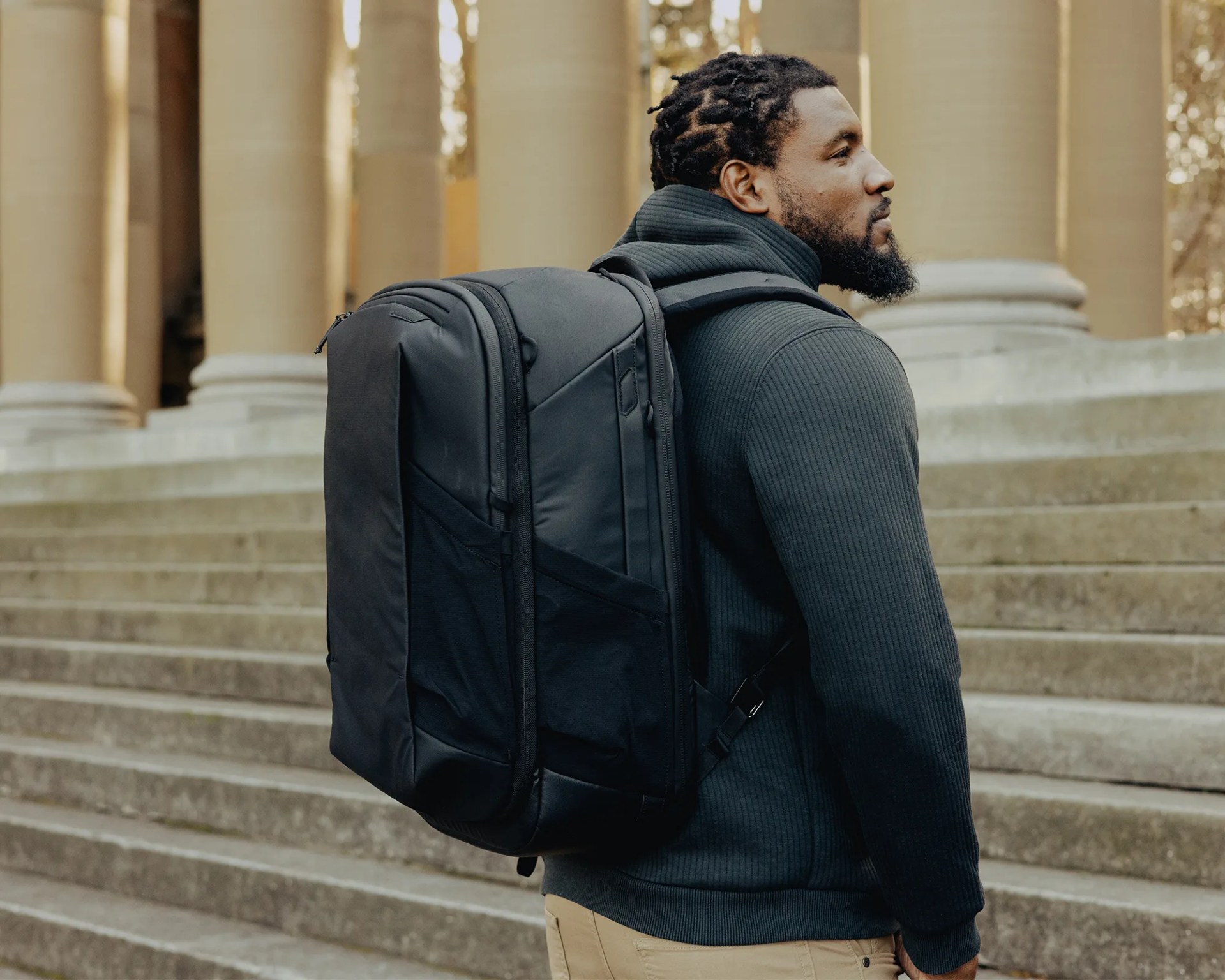 Black large backpack with multiple compartments worn by a person standing on stone steps near columns.