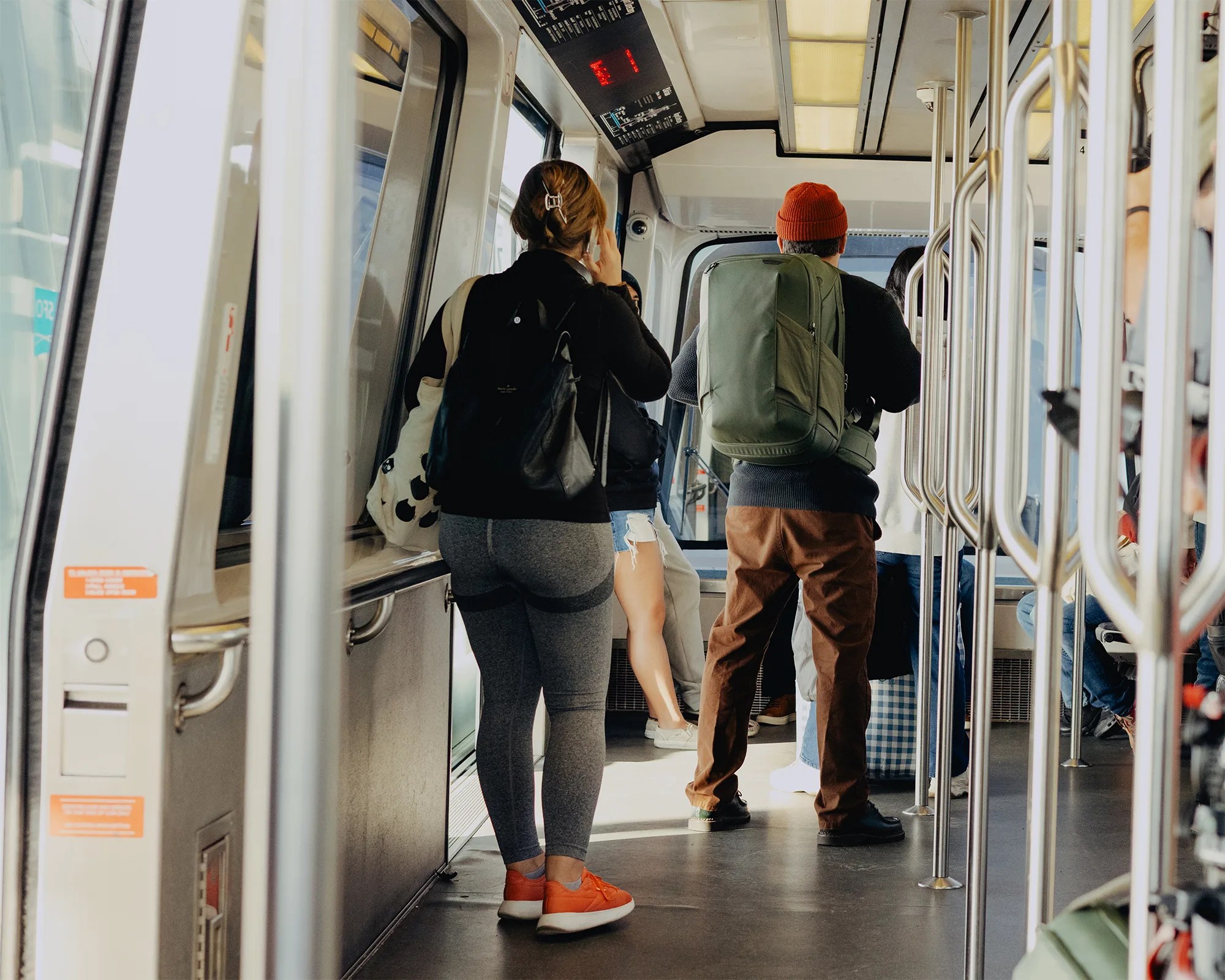 People standing inside a public transit vehicle, one wearing gray leggings and orange sneakers, another with a green backpack and red beanie.