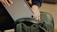 Person placing a silver laptop into an open dark green backpack with padded compartments.